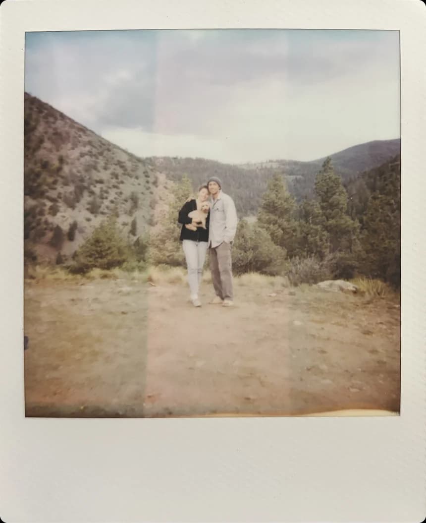Two people and a small dog standing on a dirt pull-off, pine ridges behind them.