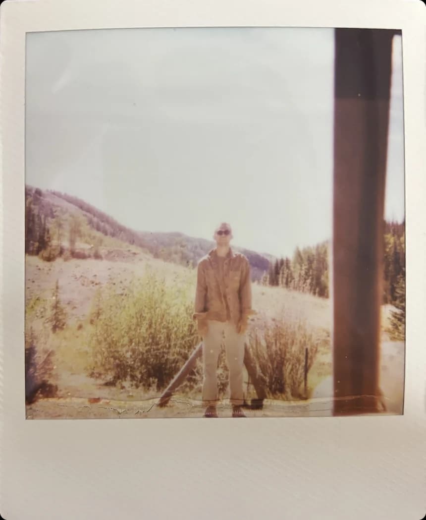 A person in a work jacket and sunglasses standing in an open mountain meadow.